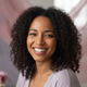 A happy woman with dark, curly hair and warm brown skin smiles, in a soft studio setting with rose and dusty lilac tones.